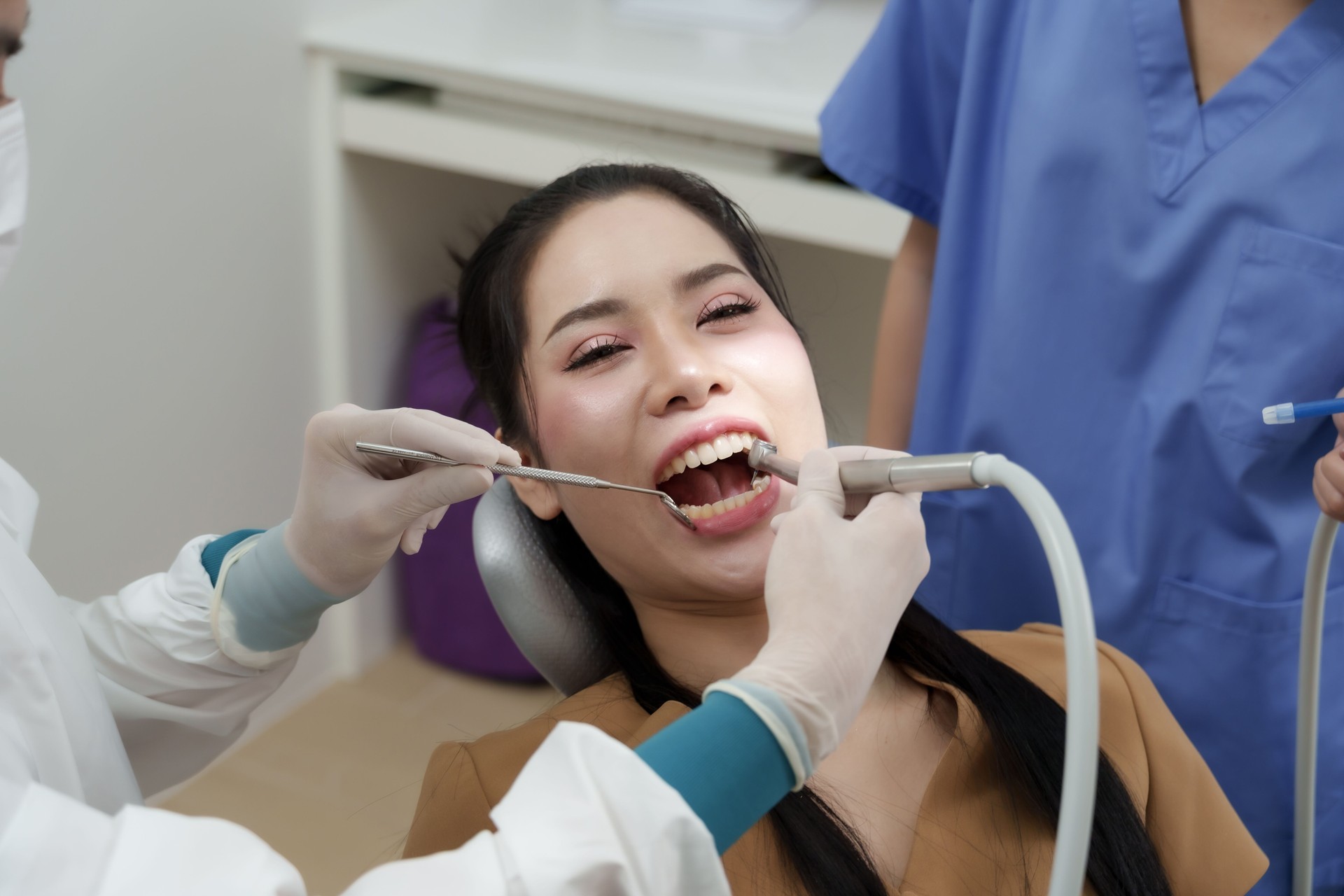 asian male dentist using suction tool and dental probe to examine teeth of smiling asian female patient while female assistant standing nearby during professional dental care session in clinic
